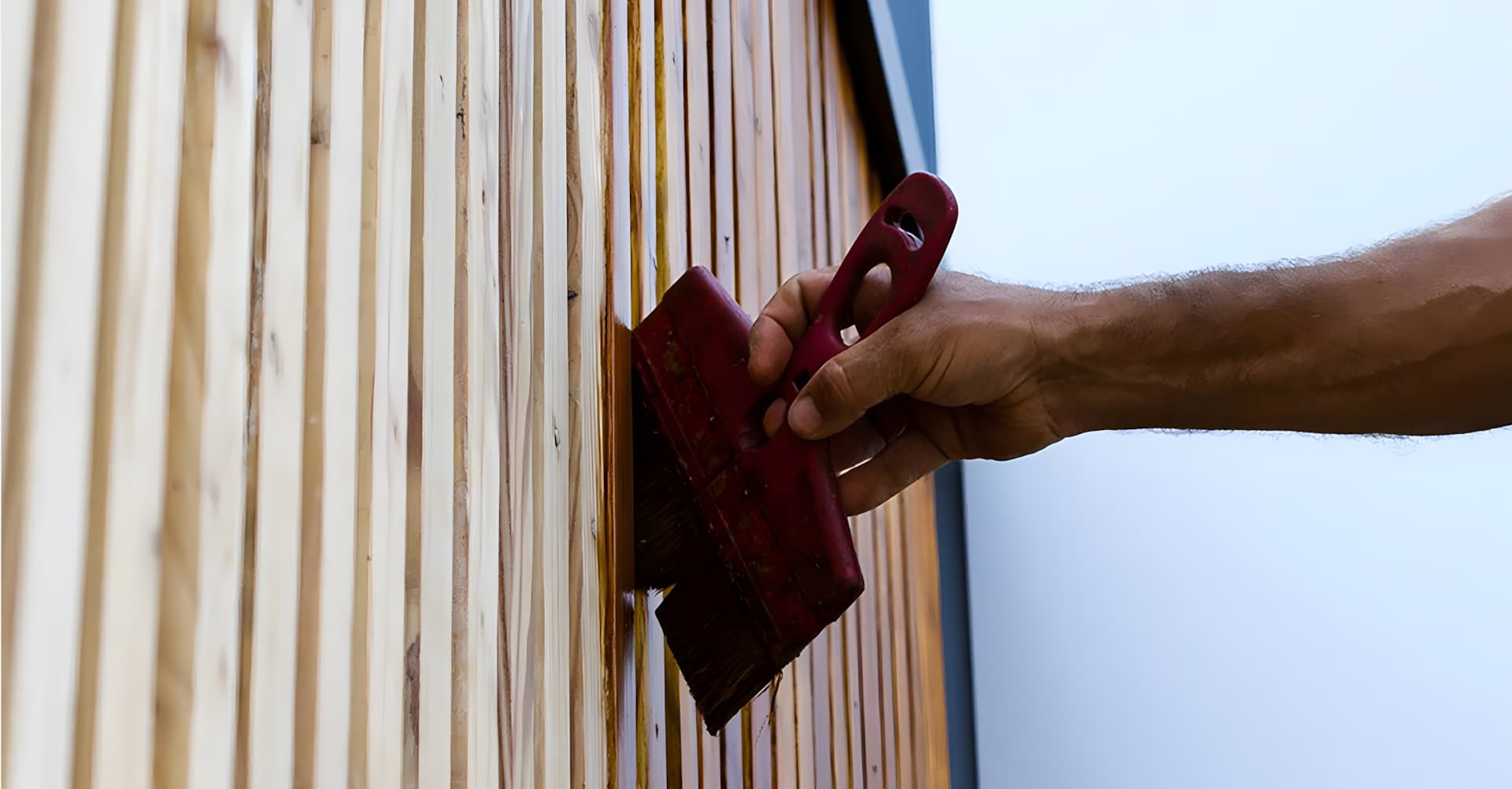 Une main applique au pinceau une peinture spéciale entretien extérieur pour bardage bois.